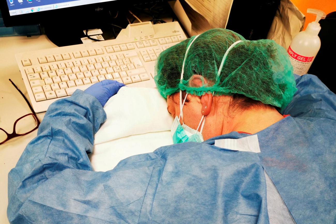 The image shows a nurse still waring a mask, gown, gloves and other protective gear sacked out with her head on the desk in front of a computer, apparently asleep. 