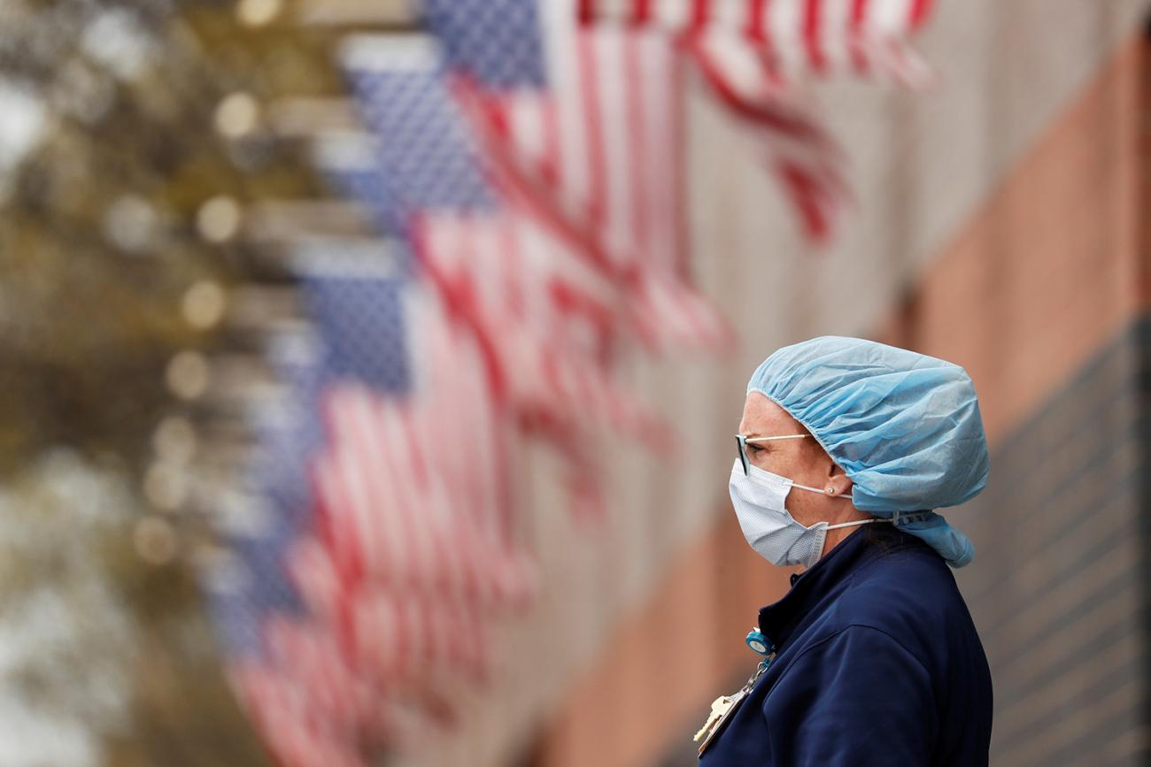 The photo shows a nurse wearing personal protective equipment leaning against a fence that is topped by many American flags. 