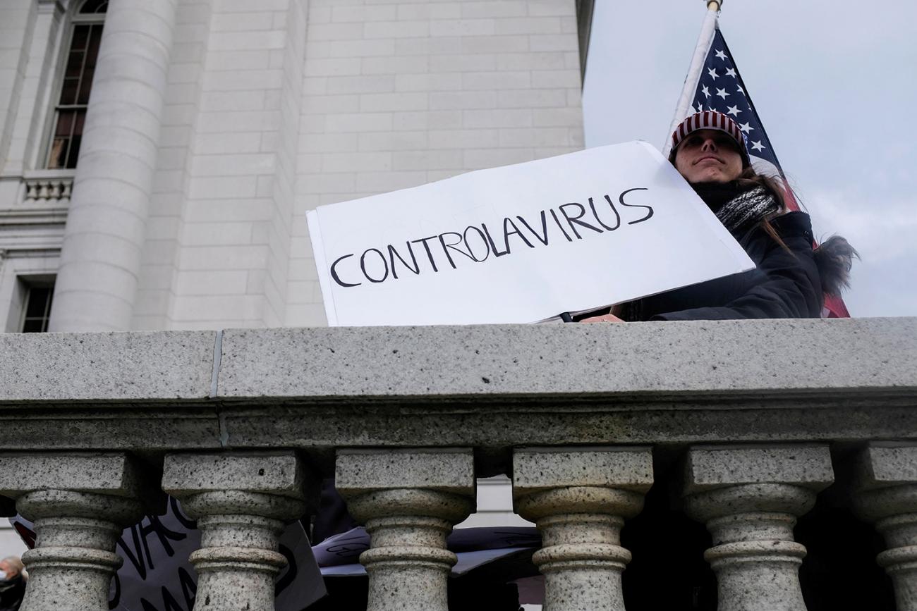 The protestor can be seen against the backdrop of the capitol building and an American flag holding a handmade sign that reads simply, "Controlavirus."
