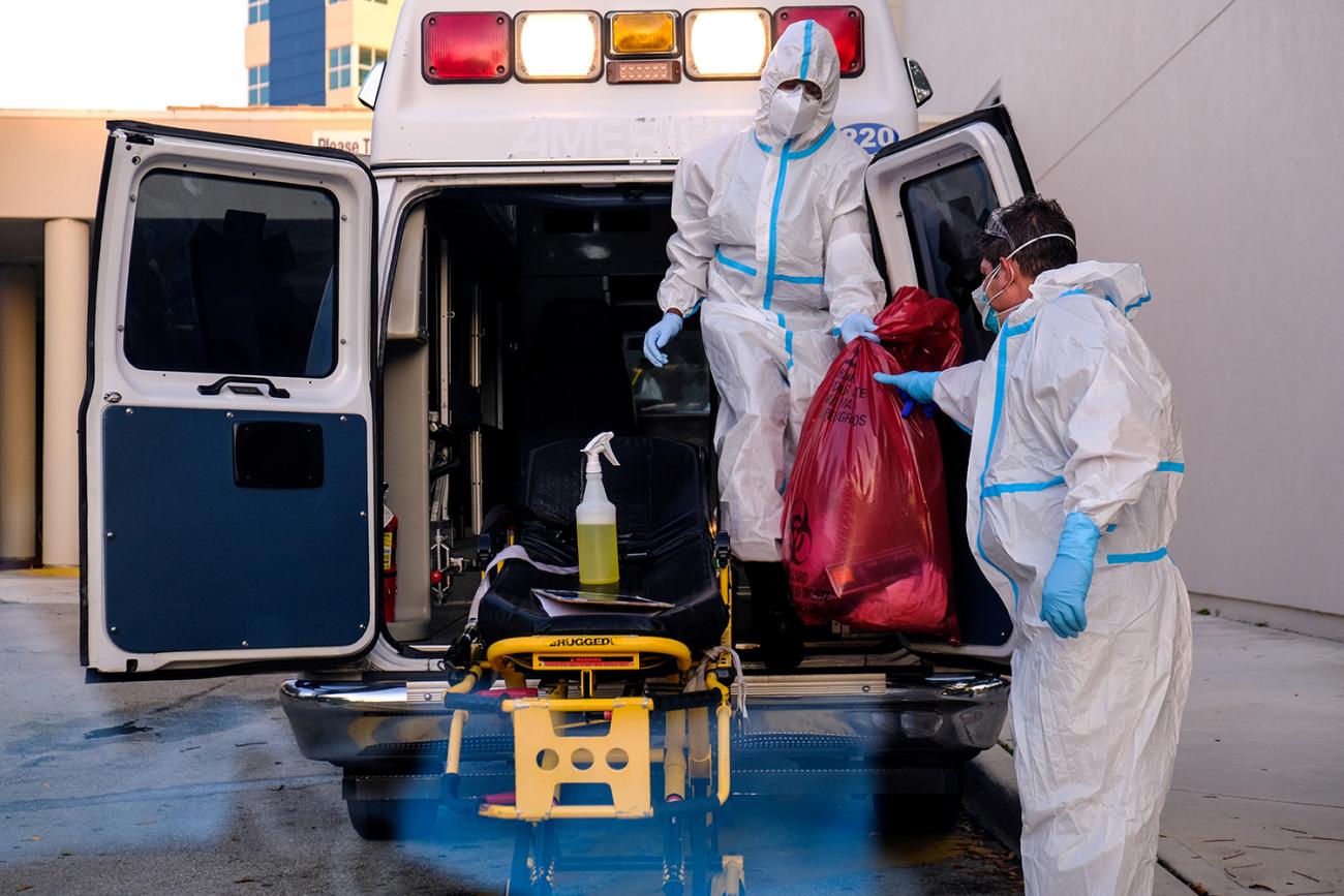 The photo shows a pair of health workers coming out of the back of an ambulance with an orange bag. 