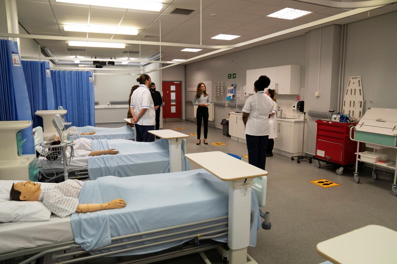 The photo shows the duchess in a hospital ward with several people around her as she speaks. In several of the beds are mannekins. 
