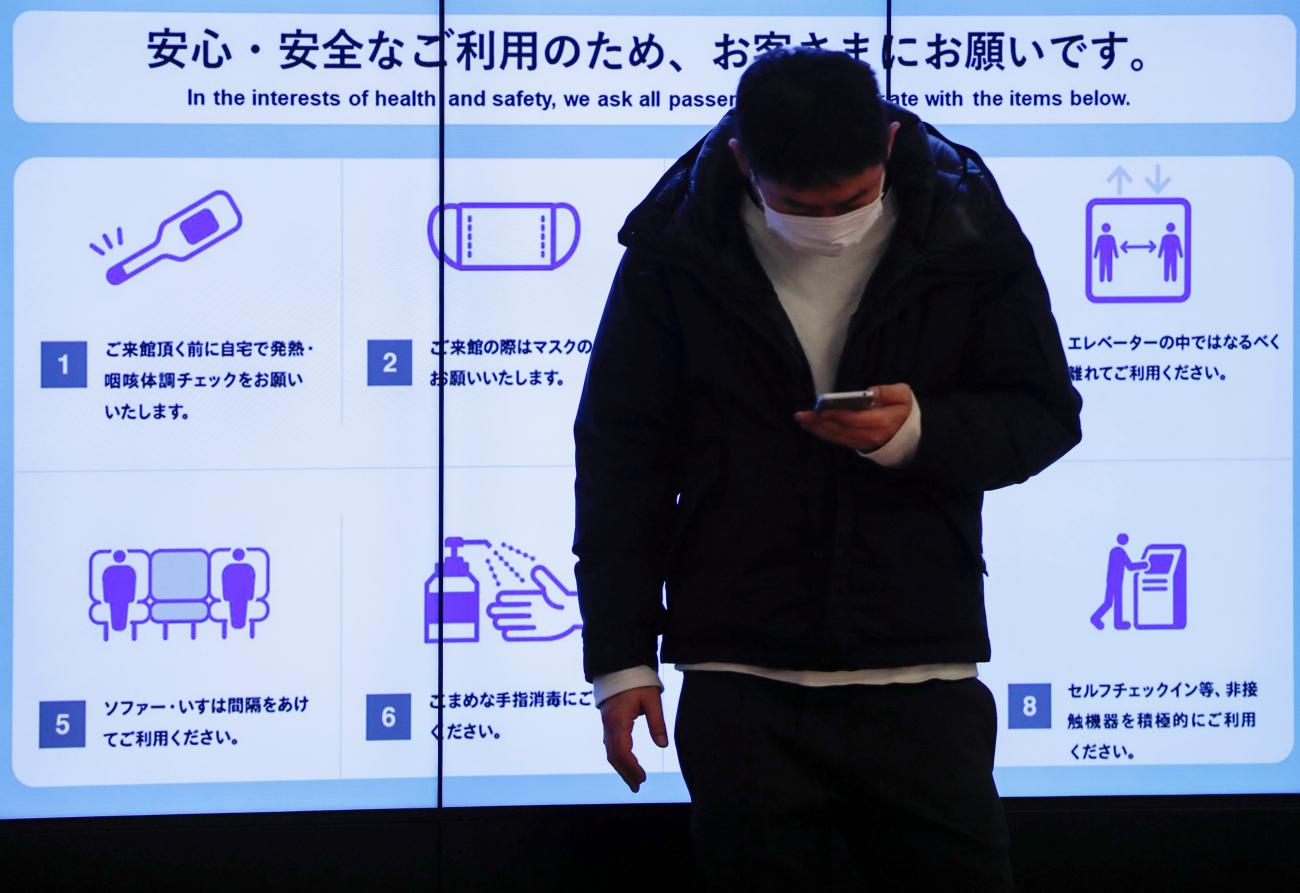 A man wearing a protective face mask stands in front of an electric screen displaying initiatives to prevent infections at Tokyo International Airport in Japan on Japan December 28, 2020.
