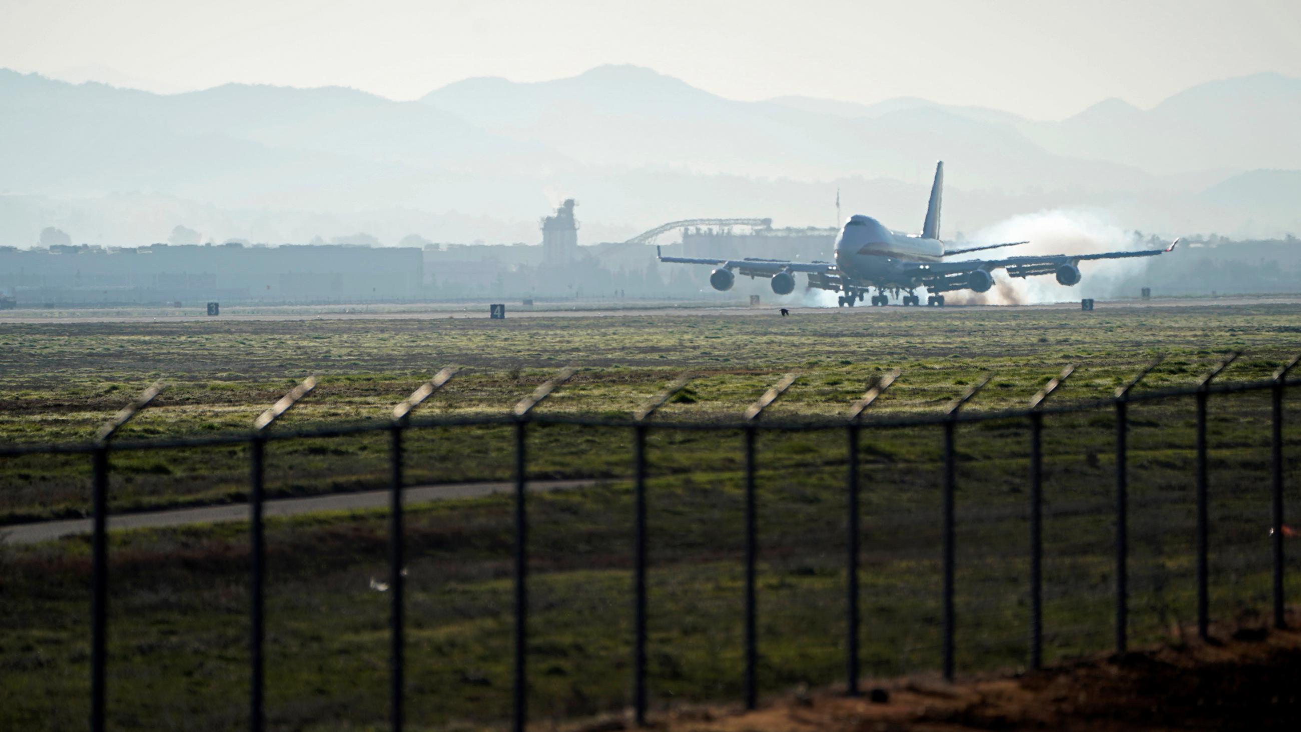 Photo shows a plane touching down in the distance from behind a security fence. 