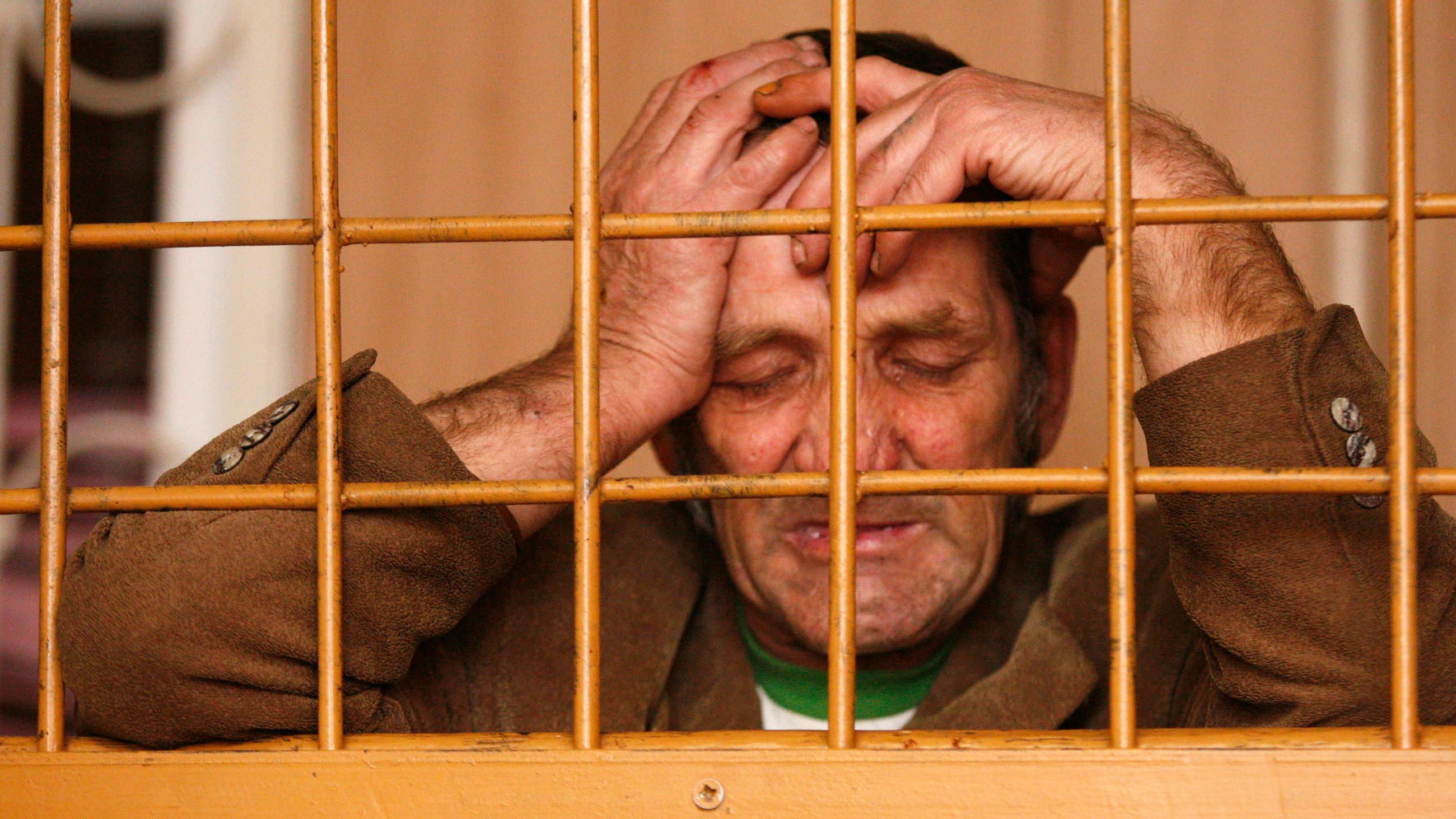 Photo shows a drunk man, 46–60 years old, eyes closed with his hand on head seen through the bars of a holding pen. 