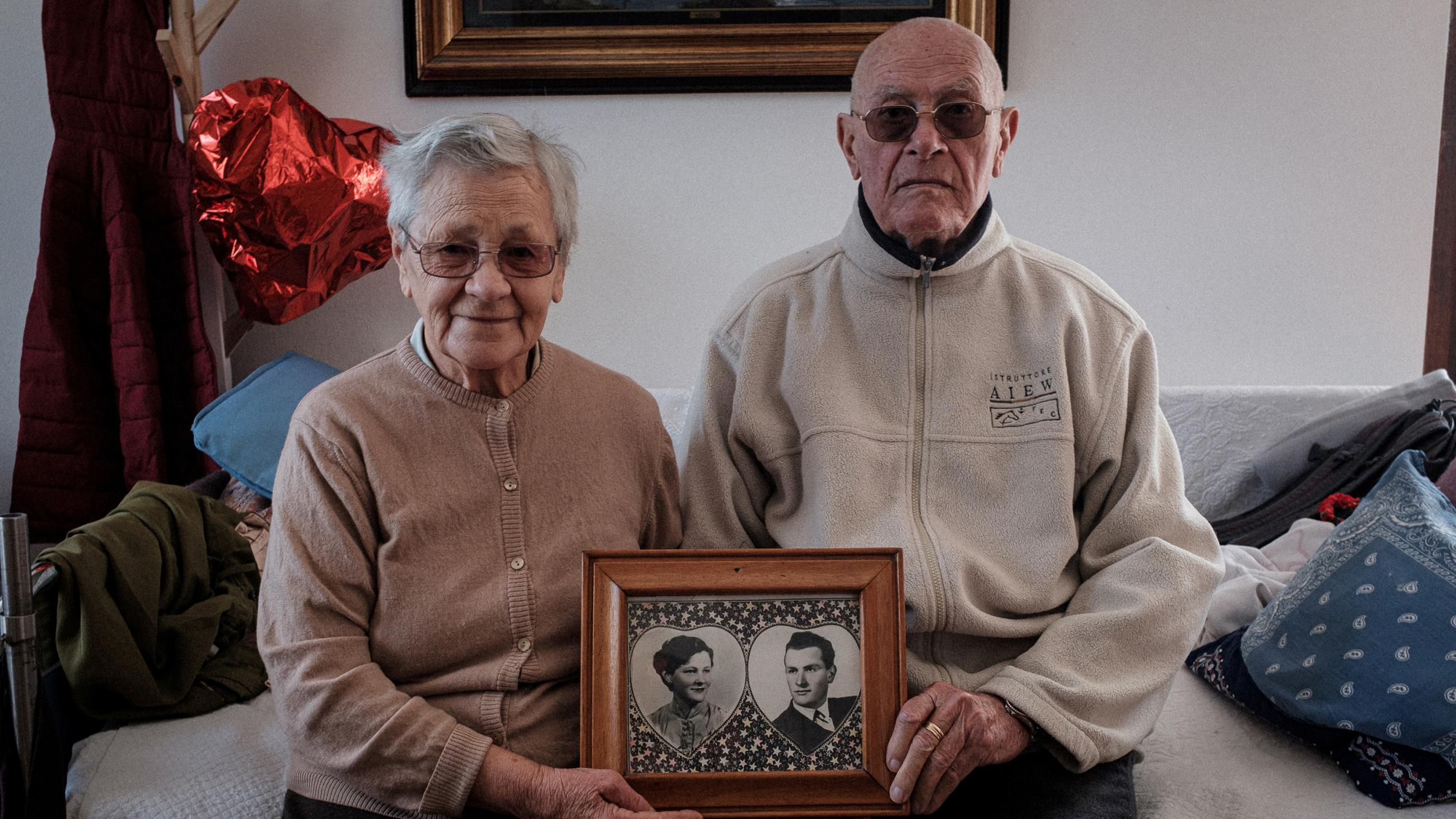 Picture shows the elderly couple sitting together holding a picture of the two of them taken many years before. They are a cute couple. 