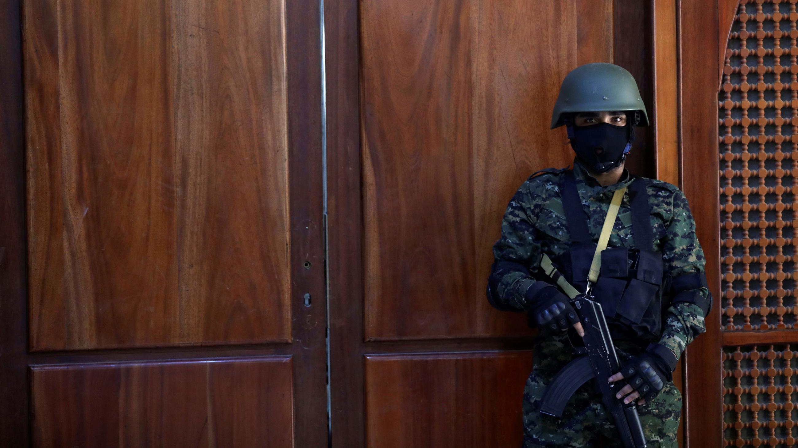 The picture shows a guard with a machine gun standing in front of a wooden door. His mask is black, the door is red.  