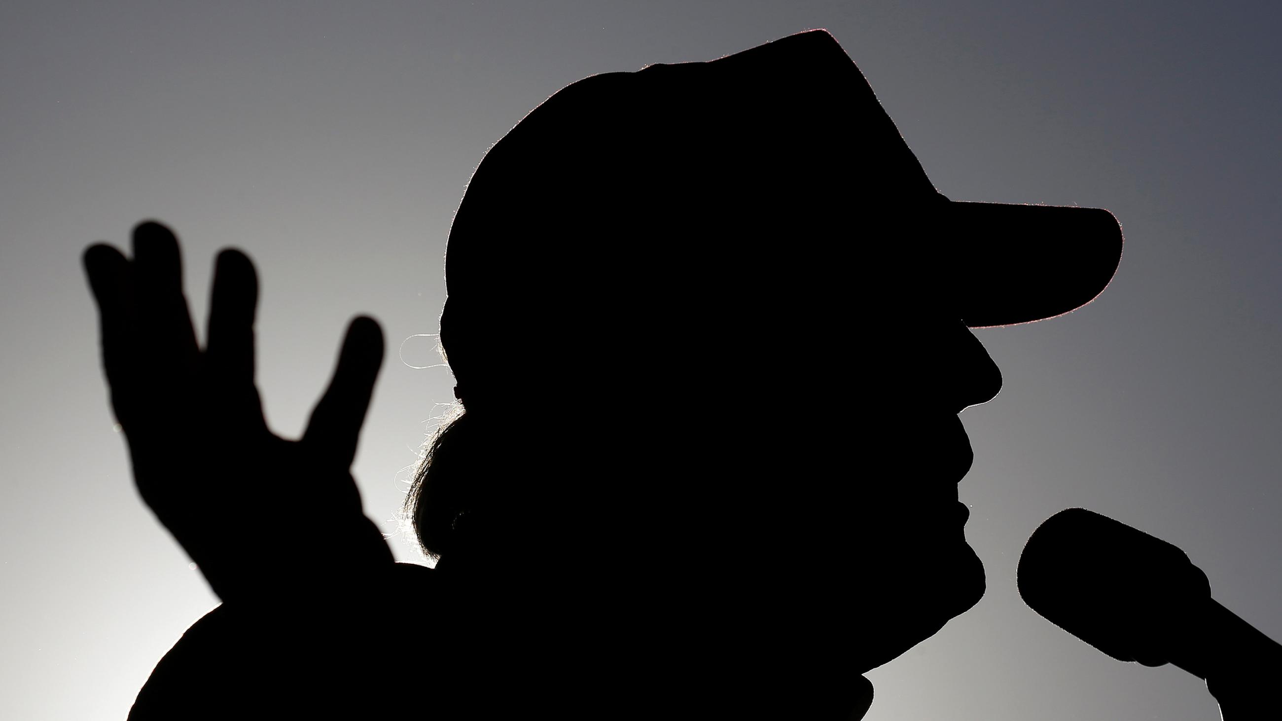 The photo shows a silhouette of the candidate speaking. he is wearing a hat and gesturing with his hand. 