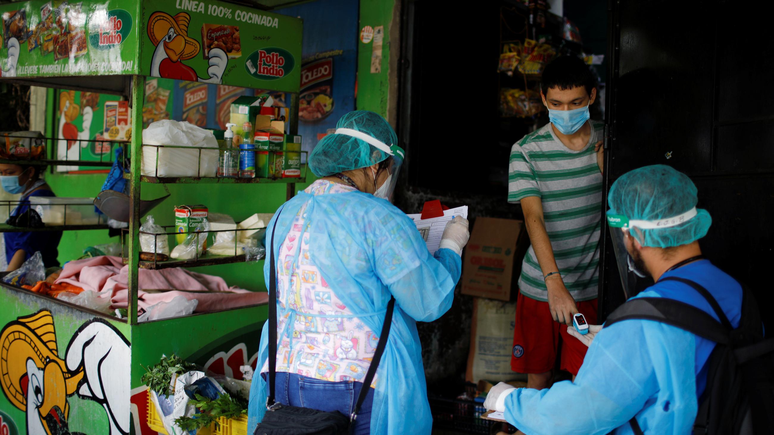 The photo shows two health workers interviewing a young person at the entrance of a small market. 