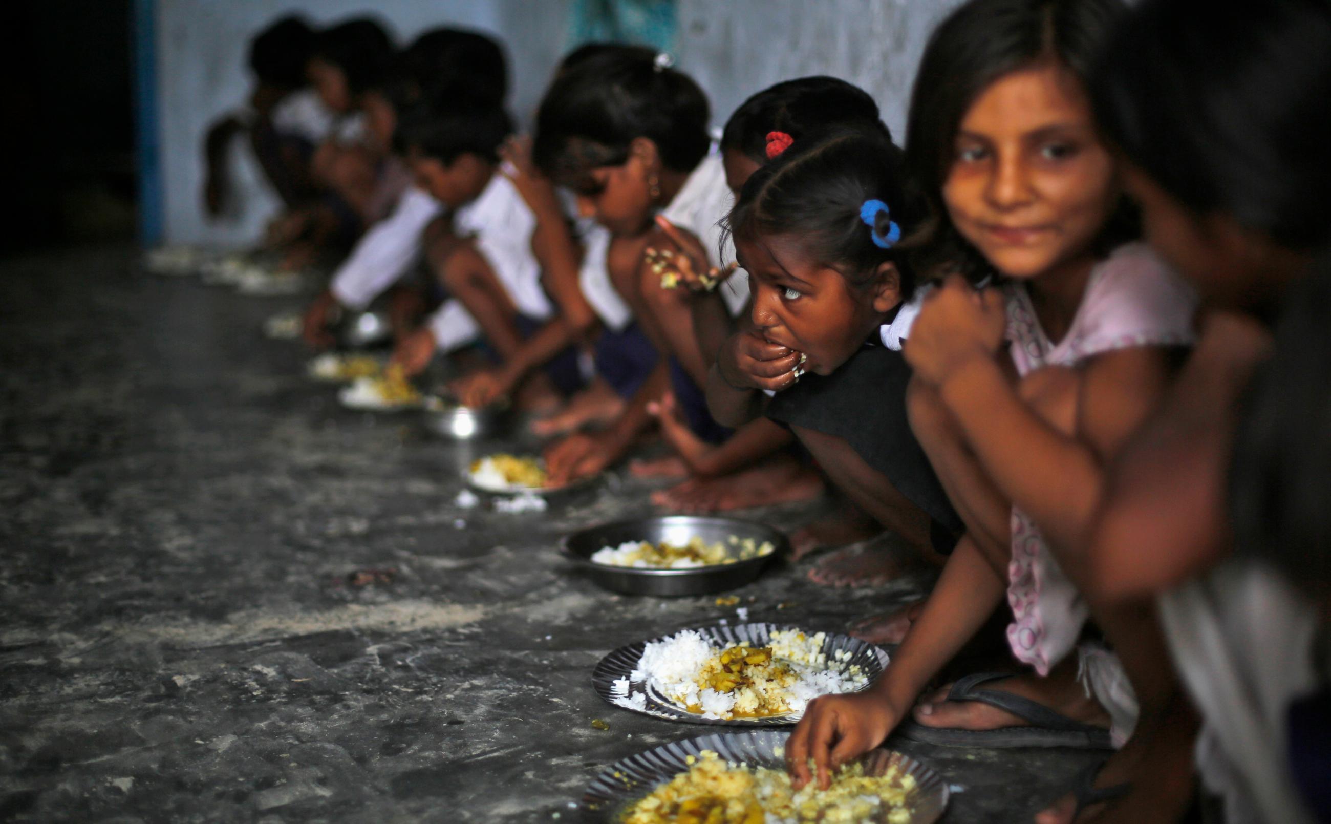 The photo shows a class of young children lined up against a wall, seated, and eating plates of rice and curry with their hands. 