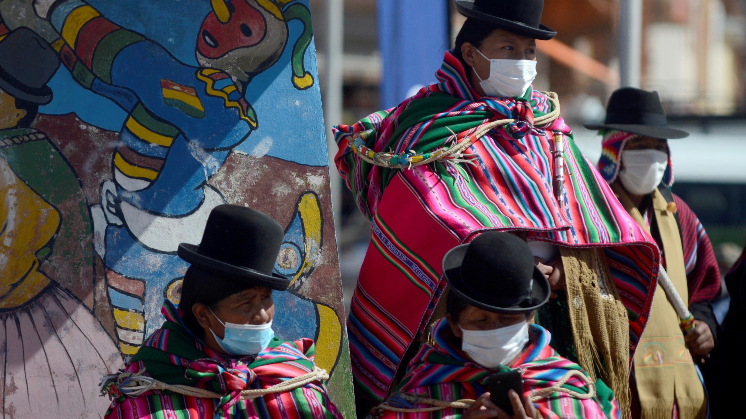 Bolivian women in traditional dress stand in front of a brightly colored mural near a site where people receive COVID-19 vaccines in Desaguadero, Bolivia.