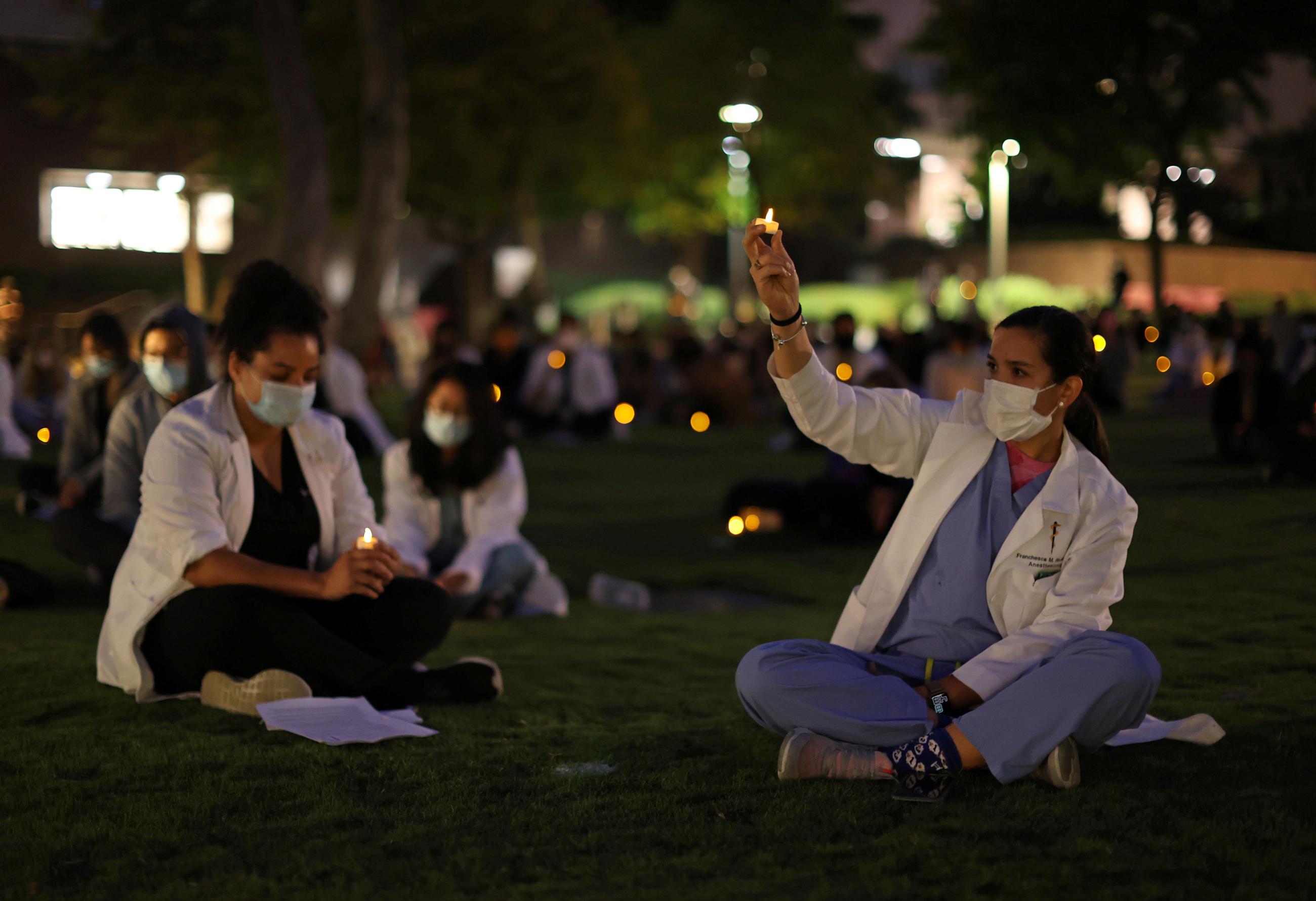 Healthcare workers in labcoats sit on a lawn holding lit candles at a vigil against systemic racism and police brutality