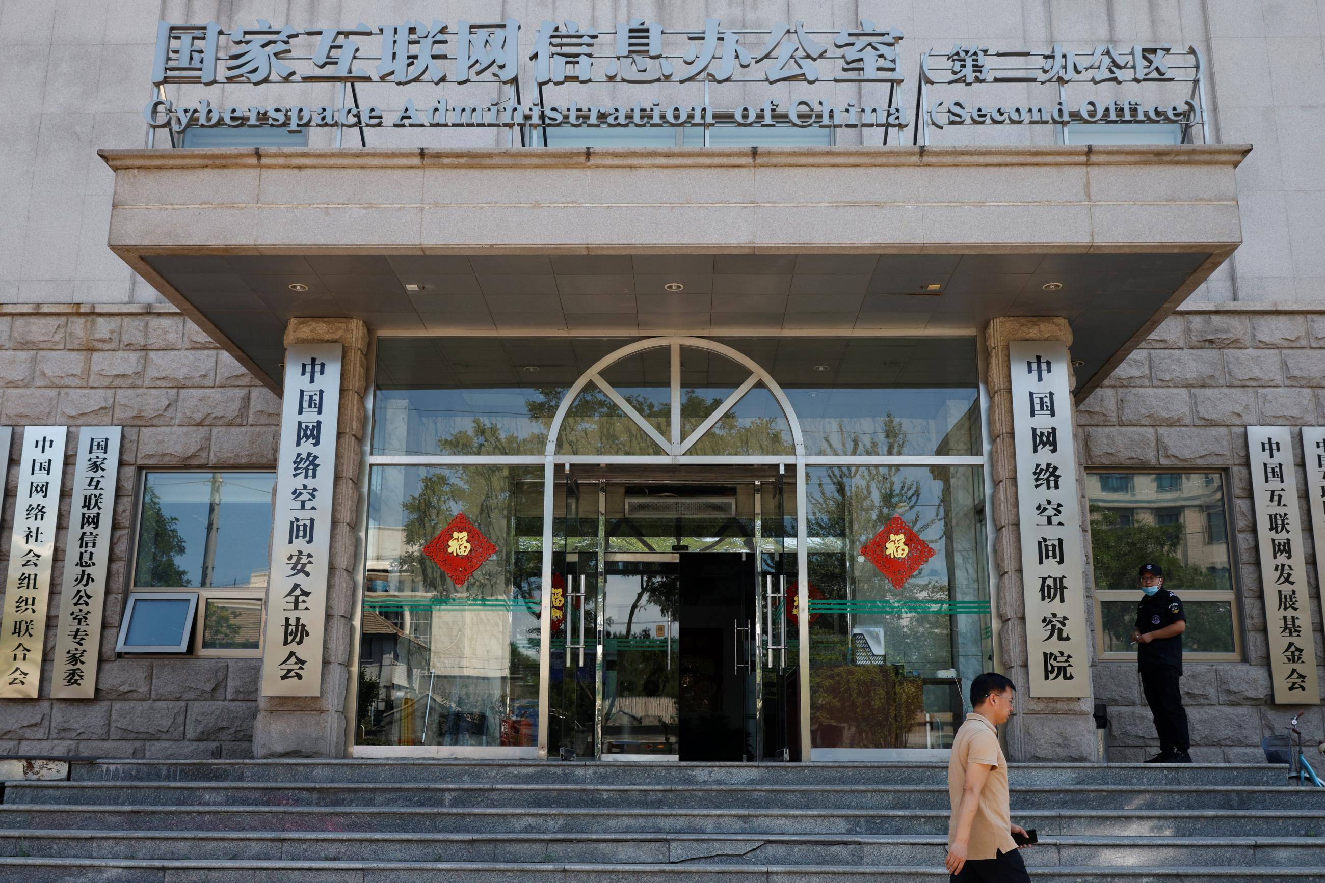 a young man wearing a beige polo shit walks bask the office of the cyberspace administration of china. a cement building with a glass facade decorated with two red lanterns on either side of the doorway