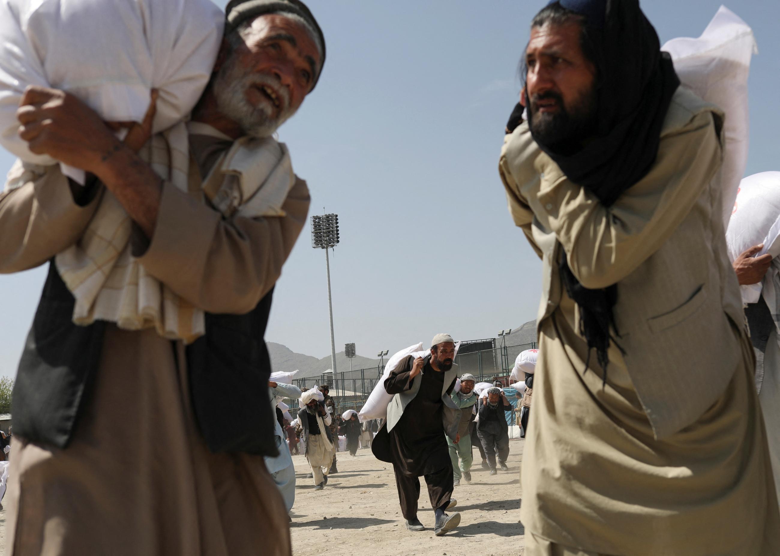 In the foreground of the photo, two men with beards carry large sacks of rice over their shoulders, while other men carry sacks in the background. The rice donations are part of a humanitarian aid effort, sent by China to Afghanistan, at a distribution center in Kabul, Afghanistan, on April 7, 2022. 