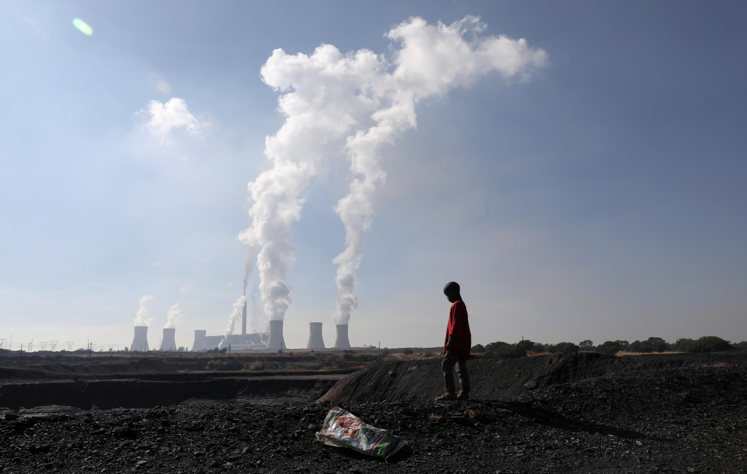 A child collecting chunks of coal looks on at a colliery while smoke rises from the Duvha coal-based power station.