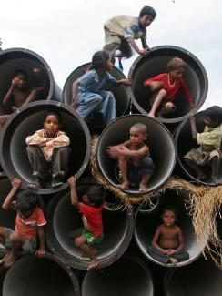 Picture shows several rows of large, concrete sewer pipes stacked on top of each other with their fat openings facing the camera. Each is large enough to fit one child huddled at the opening. About ten children can be seen climbing about on the feature.