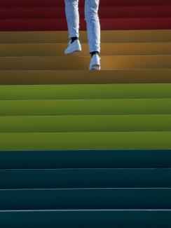 Photo shows the legs and feet of someone wearing white pants and white sneakers walking up a wide staircase that fills the frame and is brightly adorned in rainbow hues. This is a striking photo.