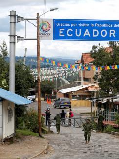 The photo shows a simple border crossing through a small town with a big sign reading "Ecuador" above the street. The crossing is closed with metal barricades, and a soldier stands out front. 
