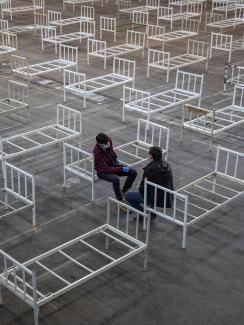 This is a striking image of two people resting on the edge of plain metal bed frames in a large space in which many, many more bed frames are placed. 