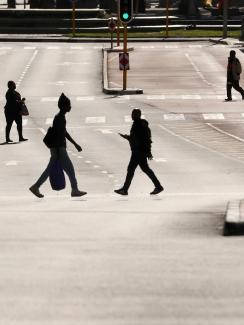This is a stunning photo showing a broad main thoroughfare that is completely bereft of cars. Four people are seen crossing the wide street, each silhouetted against the early or late sun reflecting off the asphalt pavement. 