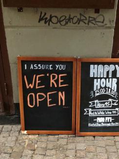 Picture Shows two side-by-side chalk placards in front of a restaurant advertising a happy hour inside. One of the signs reads, "I assure you WE'RE OPEN." 