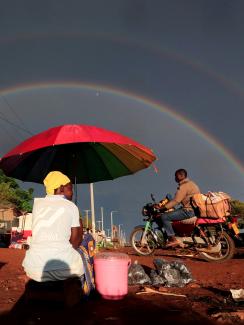 This is a spectacular photo that shows a woman under a bright red umbrella from behind as she is sitting on the side of the road greeting passer-bys with a full-double rainbow highlighting a storm-darkened sky in the distance. 