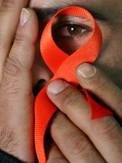 The photo shows a close-up of a man covering his face with one hand and with the other holding up a red ribbon in front of his left eye. He peers through the ribbon at the camera. 