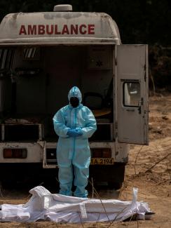 The photo shows a man dressed entirely in protective gear with his head bowed in font of an ambulance and before what appears to be a corpse wrapped in white sheets. 