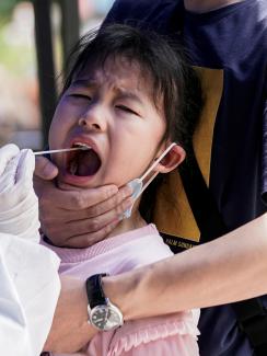 The photo shows the child being gripped by an adult while a health worker swabs a sample from her mouth. She appears to be screaming. 