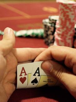 Photo shows a pair of hands holding up the corners of two cards to reveal a pair of aces. A large stack of chips is in the background. 