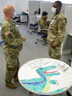 The photo shows the guard members standing around a cubicle space talking. A table in the foreground is decorated with a drawing of a dinosaur and emblazoned with the words "I eat COVID-19 for breakfast. 