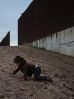 The photo shows a small child crawling on the ground near a giant border fence. 
