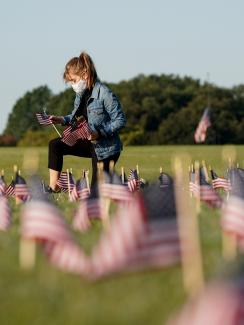 The photo shows a young woman on one knee planting a small American flag in the ground. Many more can be seen blurred and out of focus in the foreground. 