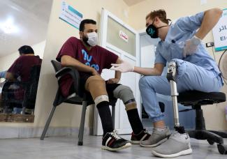 A Syrian physical therapist with a prosthetic leg, wearing blue scrubs and gesturing compassionately talks to a patient, who also has a prosthetic leg at the Al-Bab Center for prosthetics. Both men are sitting in chairs talking eye-to-eye. 