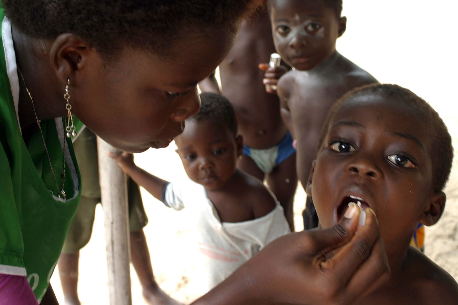 Picture shows a health care worker in a green uniform giving oral drops to a young boy who has his mouth open. Several other young children can be seen queued behind the boy.