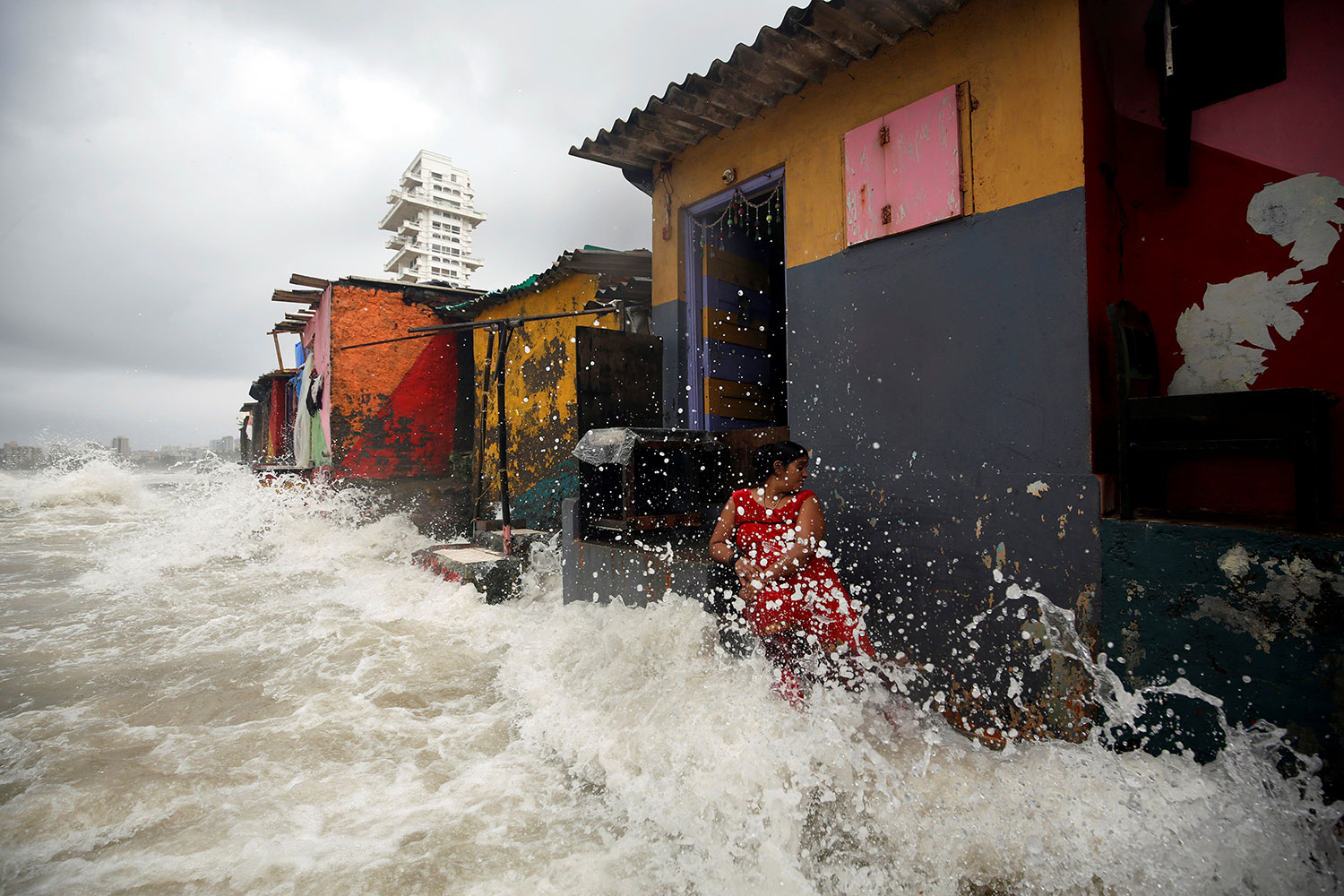 A woman is standing in front of a red, yellow, and grey slate colored tin-roof building with an open door. It is one of several structures in a row—there’s an orange one, a green one, and a yellow one. She is turning her head away from the violence of the crashing wave, which sends water shooting up all around her and nearly to the top of the building. All the area in front of the structure is covered by turbulent water. This is a frightening scene.