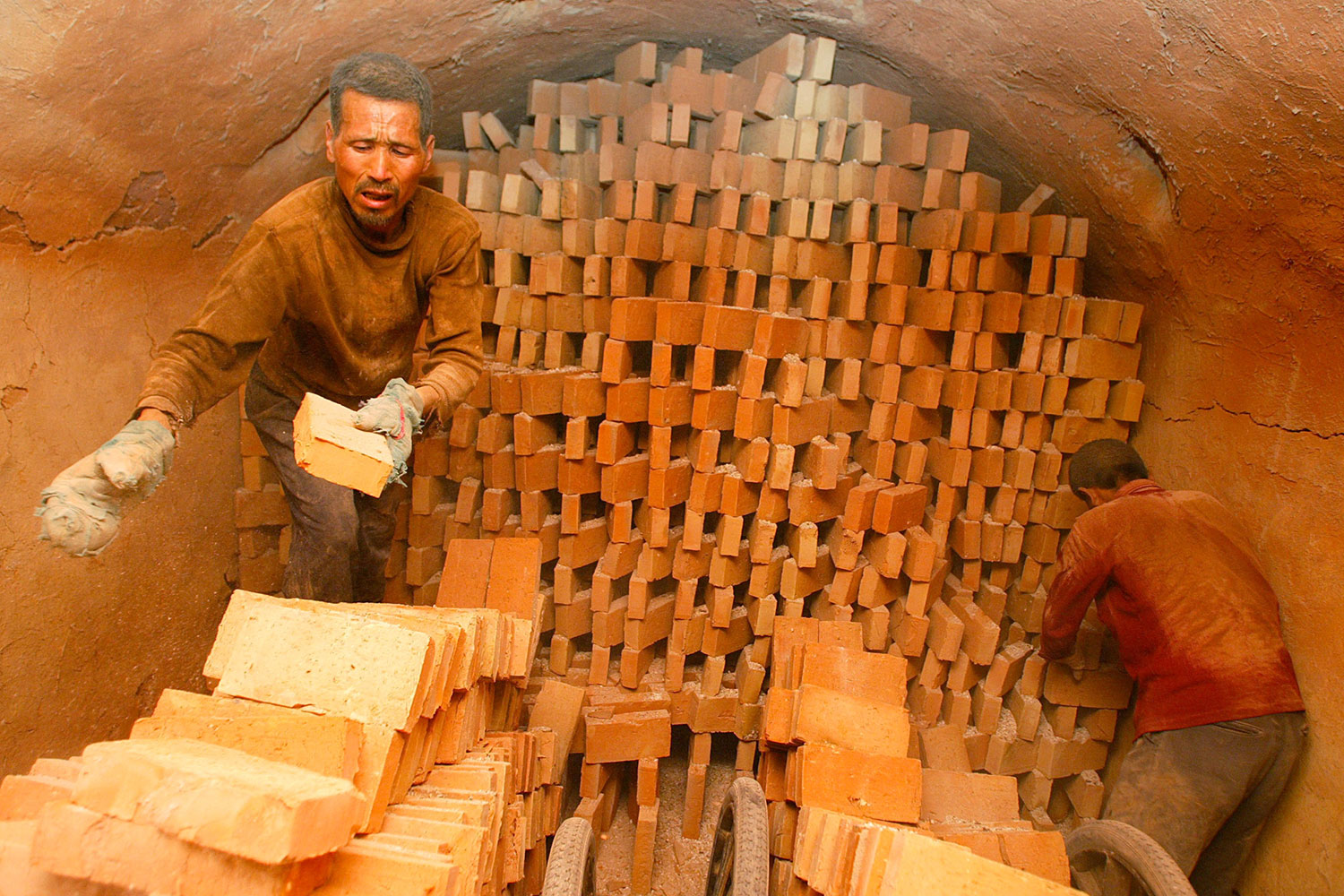 Chinese men work at a brick production plant in Xian, northwest of China's Shaanxi province on April 27, 2005. The photo shows a man pulling bricks off a stack where they were presumably kiln fired and placing them into a stack in the foreground. the overall color of the photo is brick red, suggesting that the dust is everywhere.