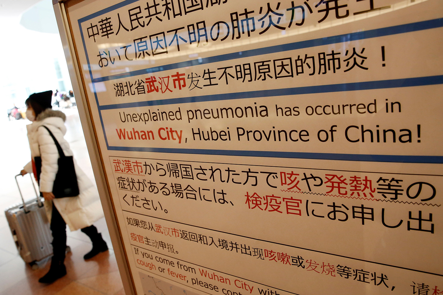 The photo shows a woman in a mask walking past a sign warning travelers in several languages.