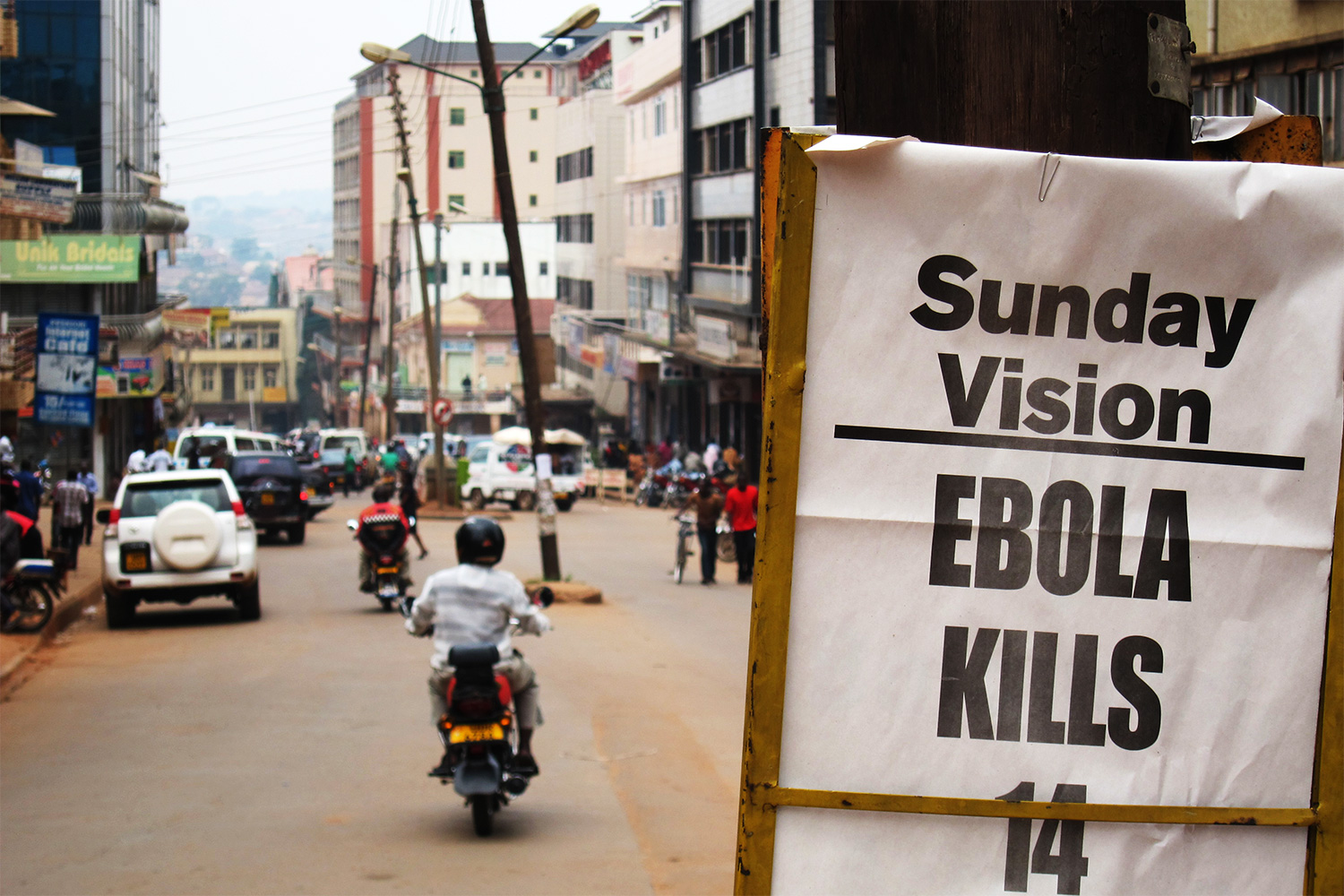 The photo shows a busy urban street with a sign announcing the Sunday paper’s headline, Sunday Vision, EBOLA KILLS 14, Sunday July 29, 2012.