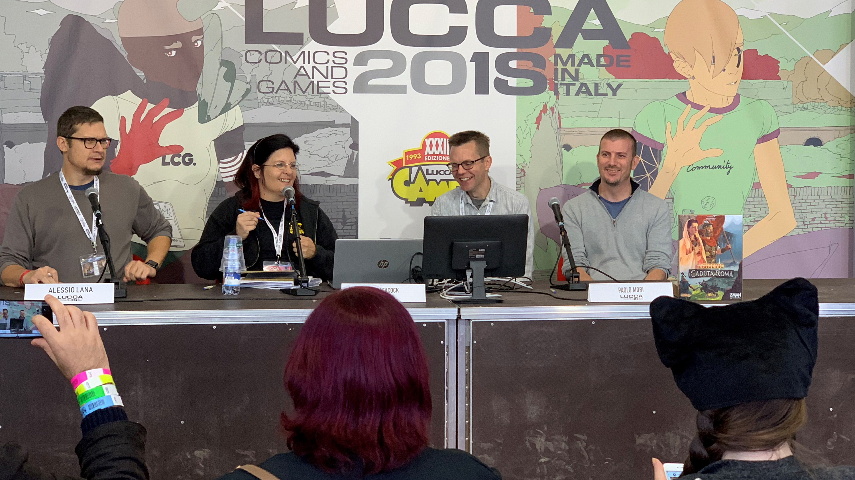 Photo shows four people on the panel, all laughing. In the foreground are three audience members with their backs to the camera—one taking a cell phone photo and the other two in costume.
