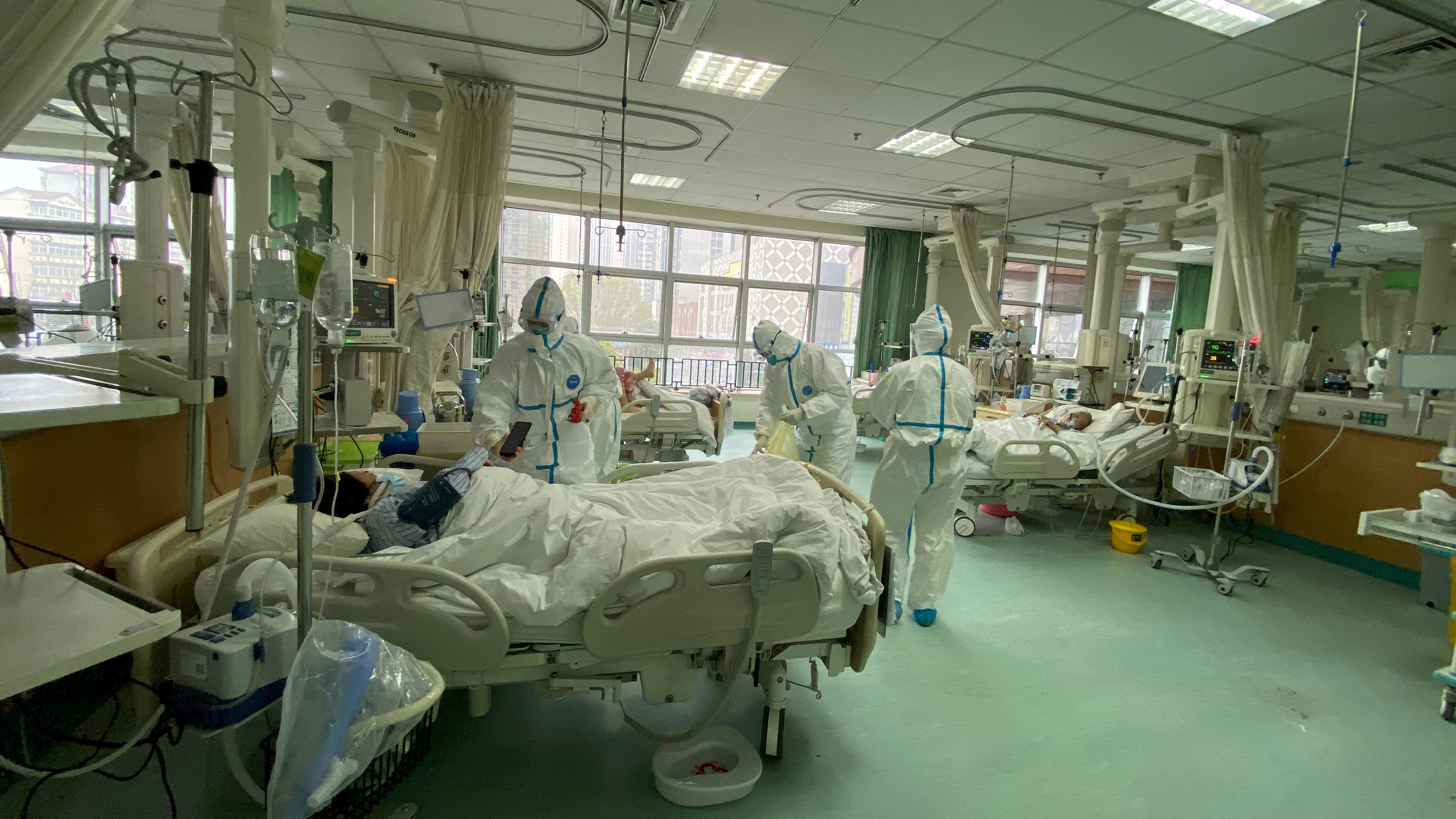 The picture shows a modern hospital ward with a patient in bed covered in protective gear as well as several health care workers covered head-to-toe in the same.