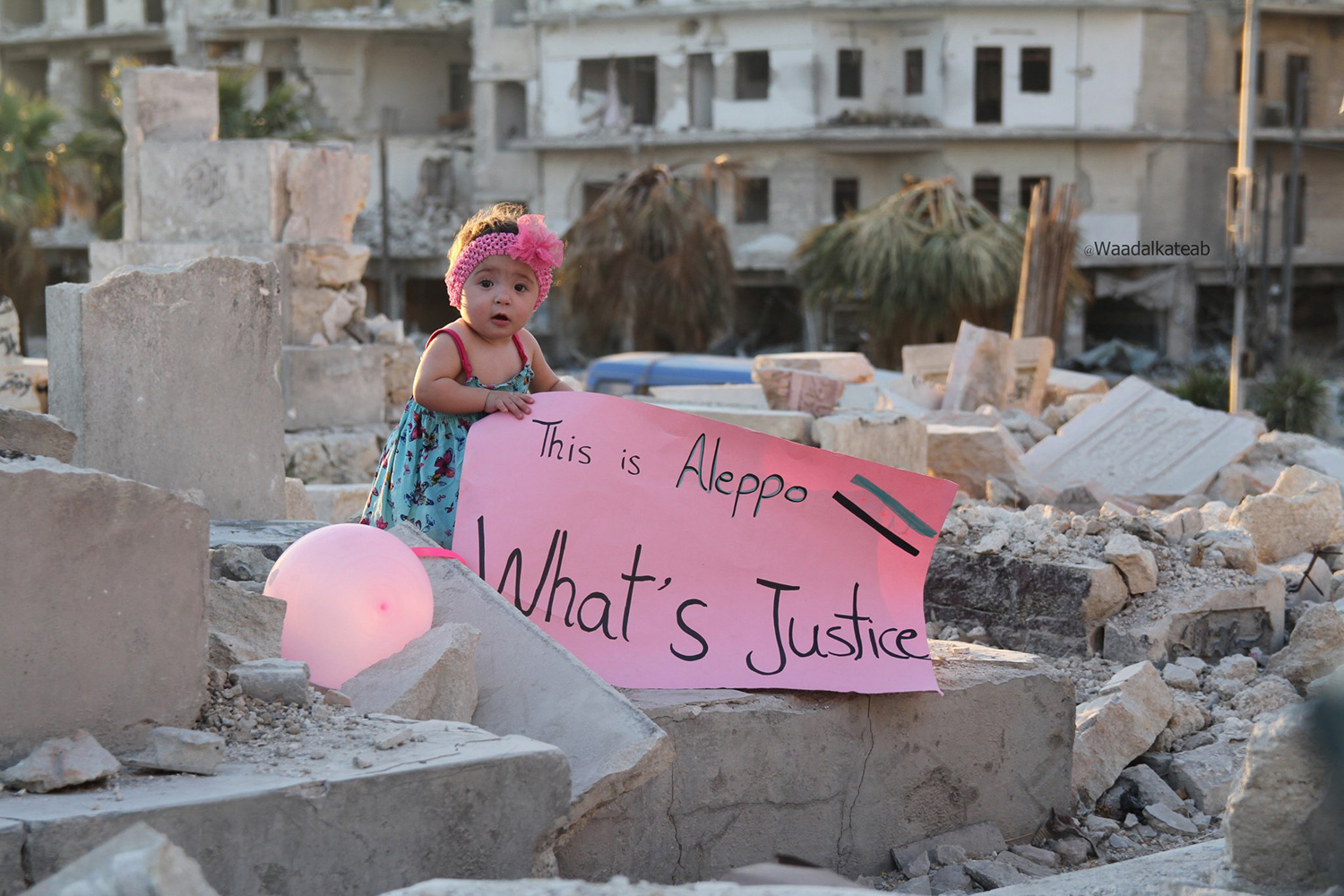 The picture shows the girl, a baby, really, standing on a pile of rubble amidst the destruction of the city holding a sign that reads, “This is Aleppo. What is Justice?” The sign is pink, and there is a pink balloon next to her, and she has a pink head band.