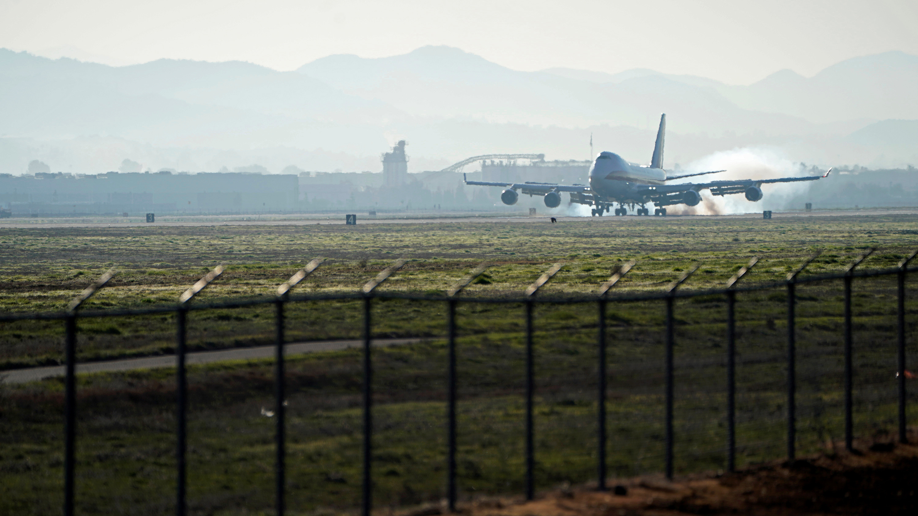 Photo shows a plane touching down in the distance from behind a security fence.