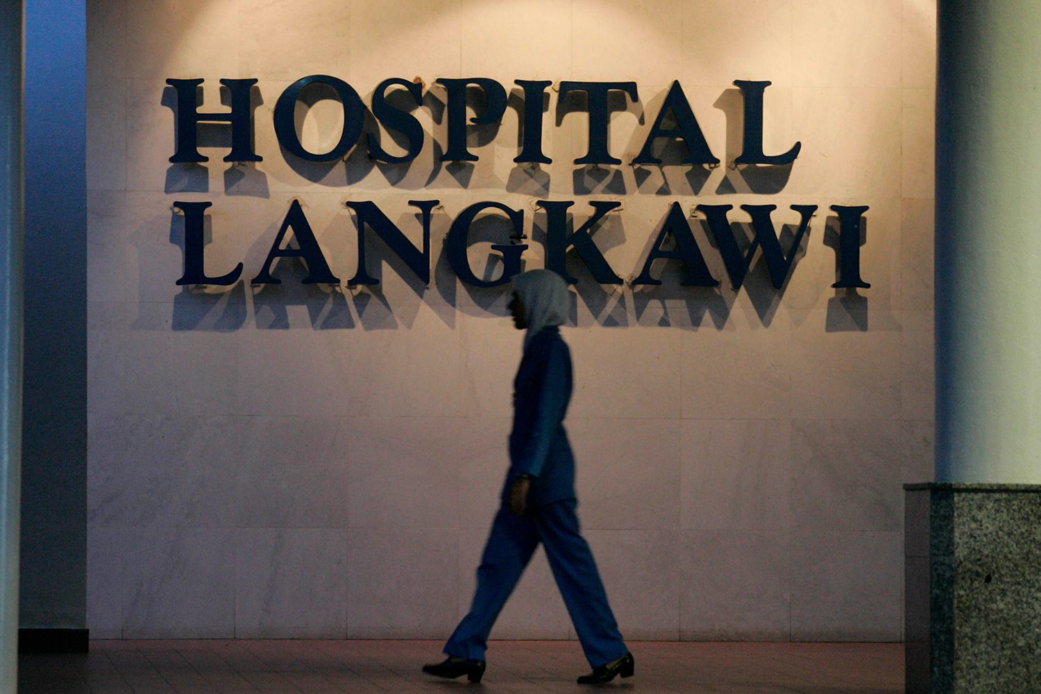 Photo shows a large hospital sign with a tall nurse walking by in front of it.