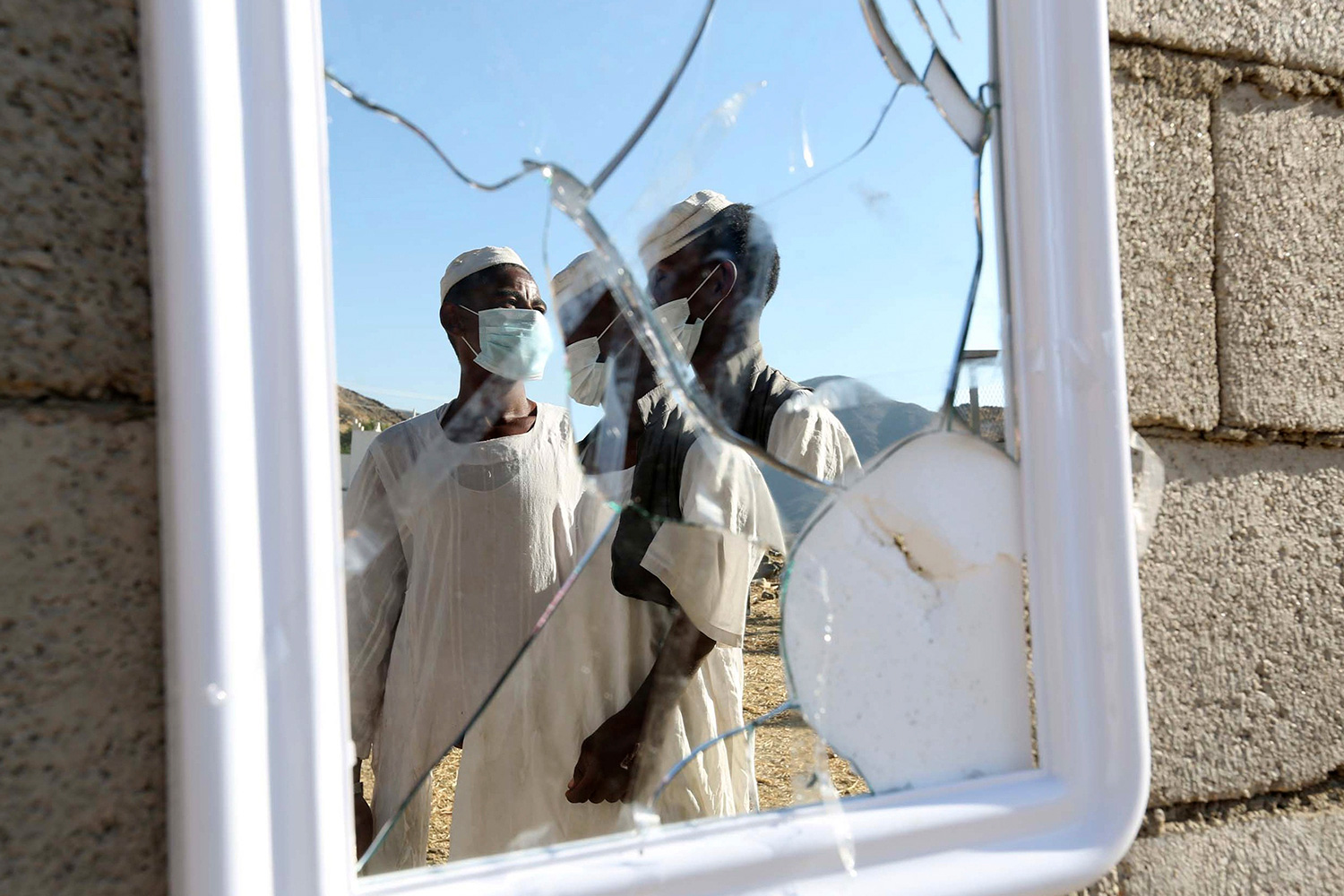 This is an artistic image depicting workers wearing medical masks who are reflected in a broken mirror posted on a cinderblock wall.