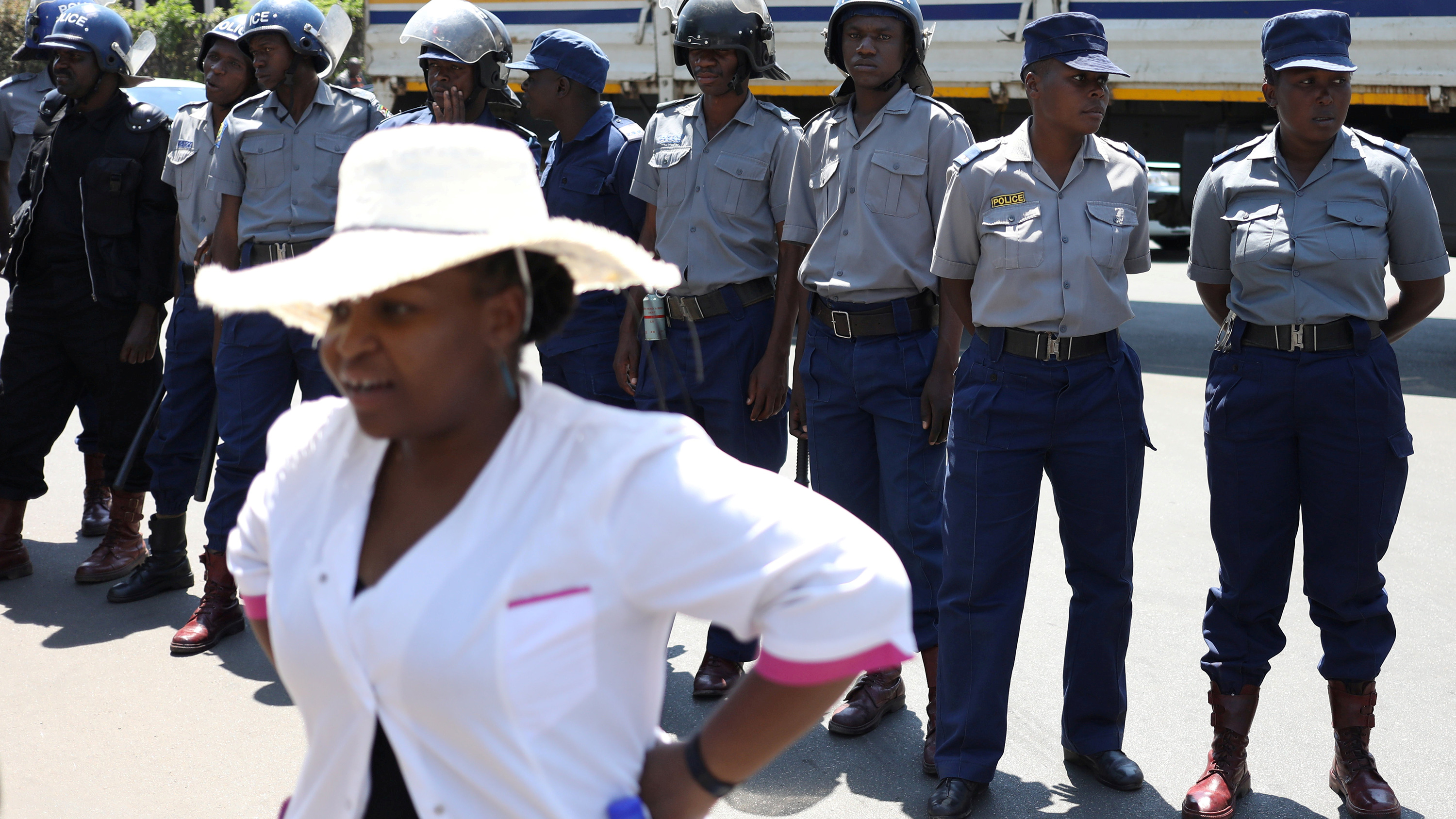 The nurse in a white shirt and wide-brimmed white hat is standing with her back facing a line of uniformed police officers, some wearing riot gear. She has her hands on her hips.