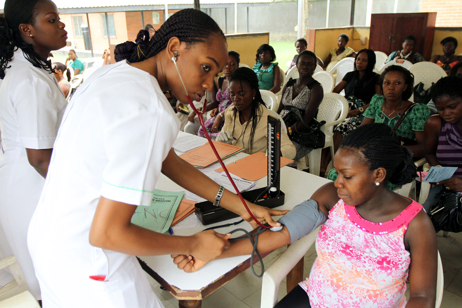 The photo shows a pregnant woman seated with a health care worker standing In front of her reading her blood pressure. Several rows of chairs behind her are filled with other women waiting.