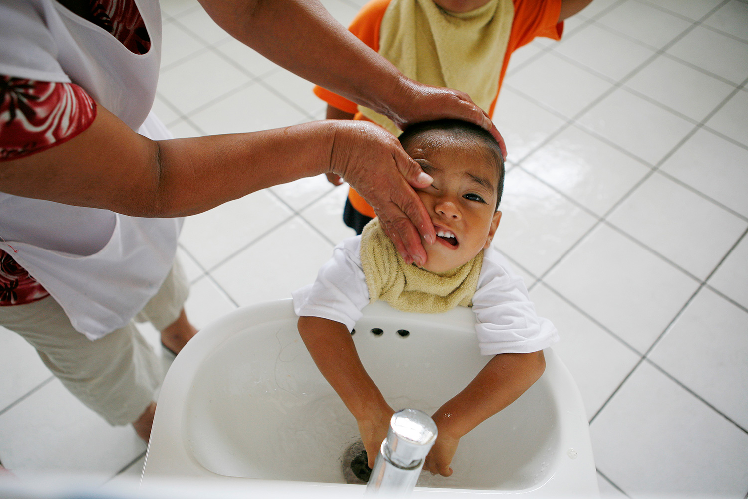 The picture shows a social worker washing the face of a child standing at the sink. He doesn’t look like he’s enjoying it.