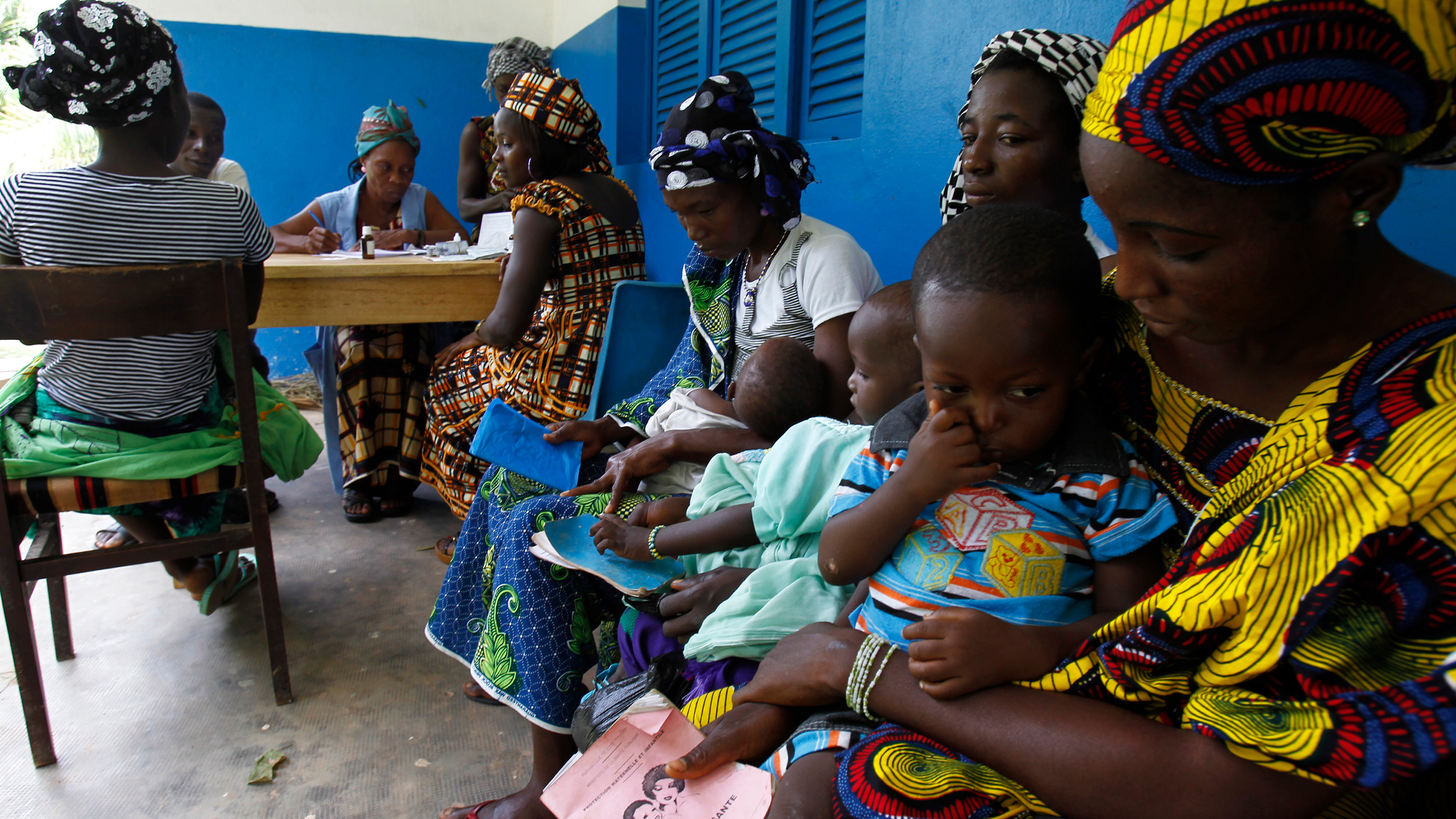 Image shows several women colorfully dressed holding young children and waiting on a bench.