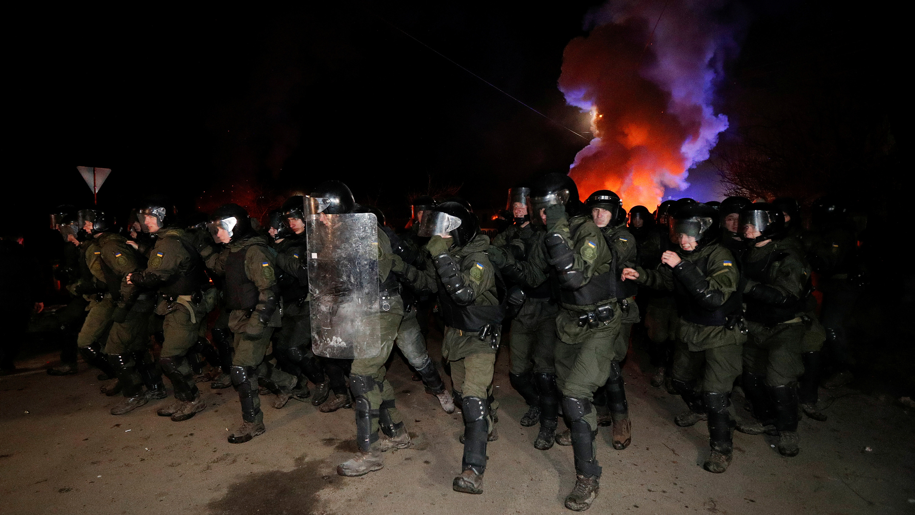 Picture shows more than a dozen officers wearing full riot gear advancing toward the camera at night. Behind them smoke rises and is illuminated by bright orange and blue light, presumably from a police vehicle off camera. This is a stunning photo.