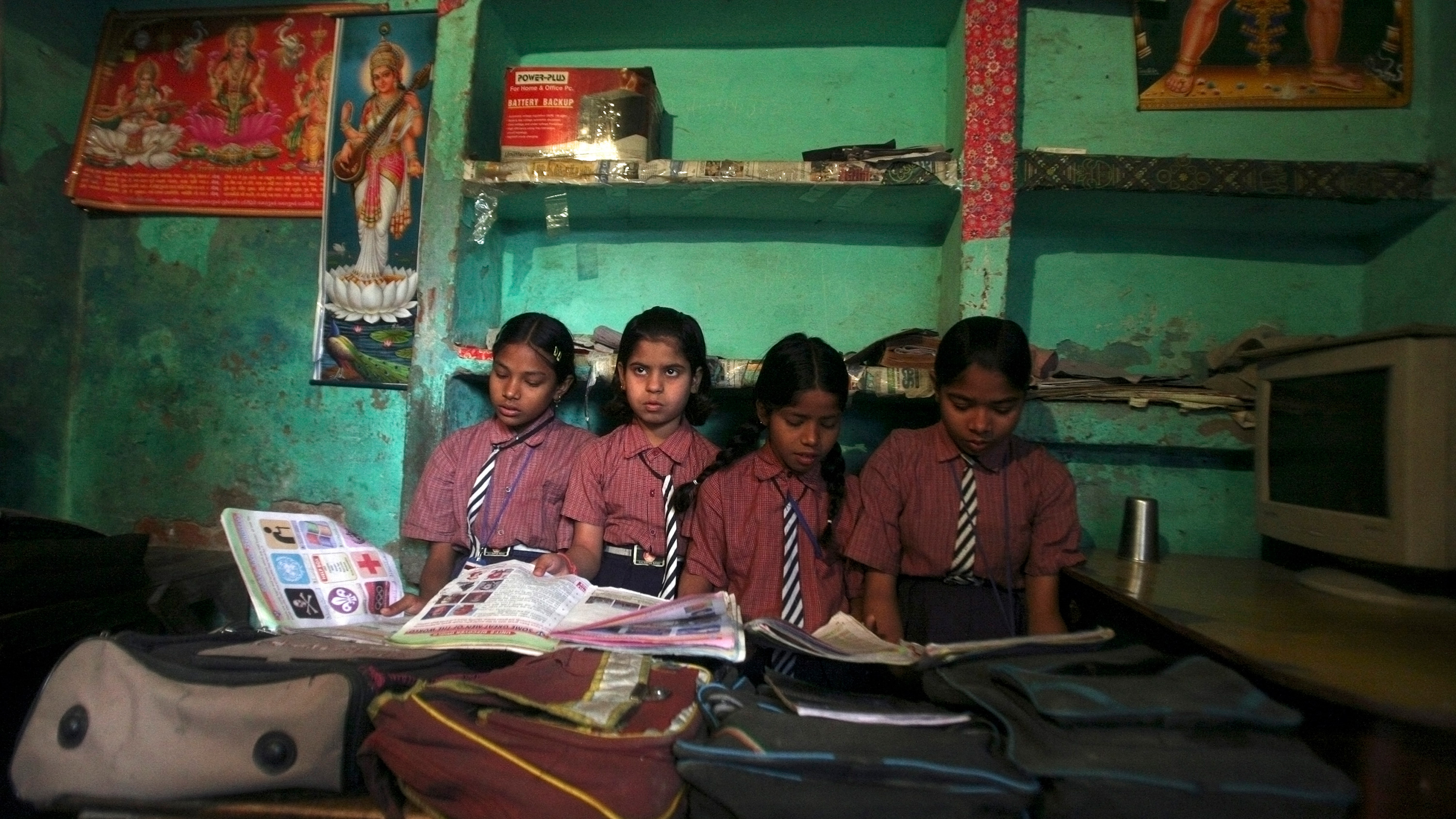 Image show four girls sitting close to each other with their heads buried in books, except one who looks up and off camera. The walls of the school are painted green.